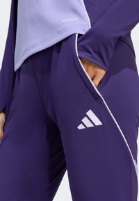 Lila träningsbyxor med vit kant och en Adidas-logotyp. Tyget har en slät textur, designat för fuktstyrning.