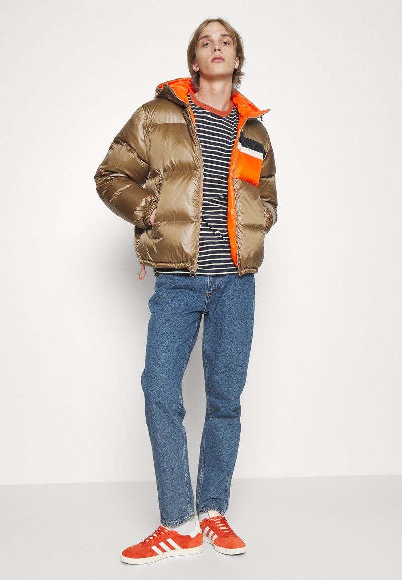 Puffjacke in metallisch Braun mit orangefarbenem Innenfutter und gestre pocket, getragen über einem schwarz-weißen gestreiften Hemd und blauen Jeans. Rote Sneakers.