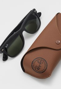 Occhiali da sole neri Ray-Ban con lenti scure accanto a un astuccio in pelle marrone. L'astuccio presenta un logo rotondo impresso e una chiusura a bottone.