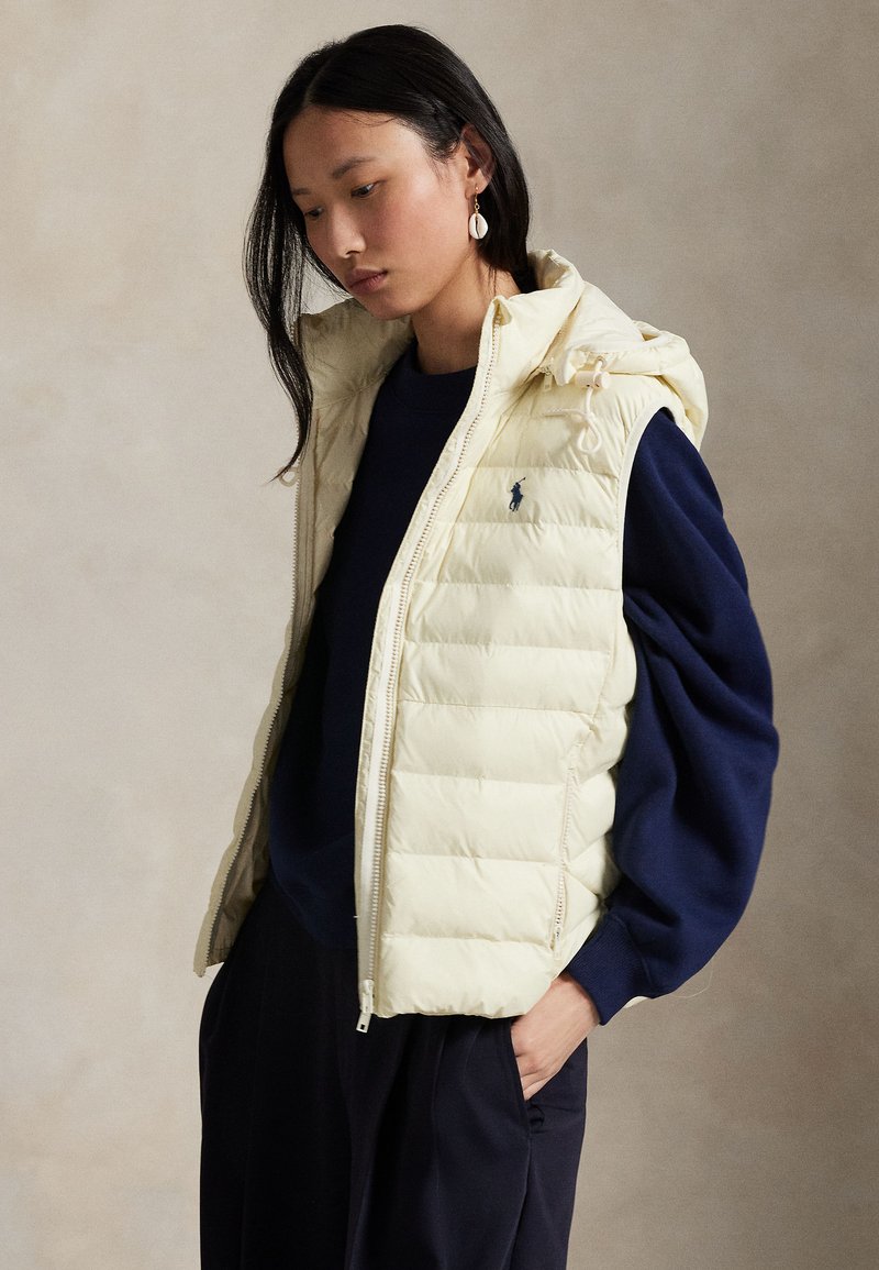 Femme aux longs cheveux foncés porte une doudoune crème sur un pull bleu marine et un pantalon noir, debout avec une main dans la poche devant un fond uni.