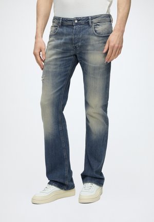 Homme portant un jean bleu délavé avec une usure subtile et des baskets blanches, debout devant un fond uni.