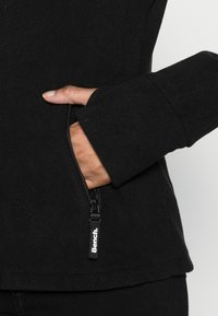 Zwarte fleecetrui met zijzak met rits, brede mouwen en een ronde zoom. De stof heeft een zachte, gestructureerde afwerking.