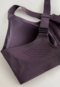 Donkerpaarse sportbeha met verstelbare bandjes en kleine ventilatiegaten op het voorpaneel, vlak gelegd op een lichte ondergrond.