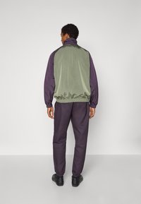 Μπουφάν αδιάβροχο με πράσινη πλάτη και μοβ μανίκια, ελαστικά μανσέτες και ζώνη στη μέση. Συνδυάζεται με παντελόνι ίδιας απόχρωσης σκούρου μοβ και μαύρα παπούτσια.