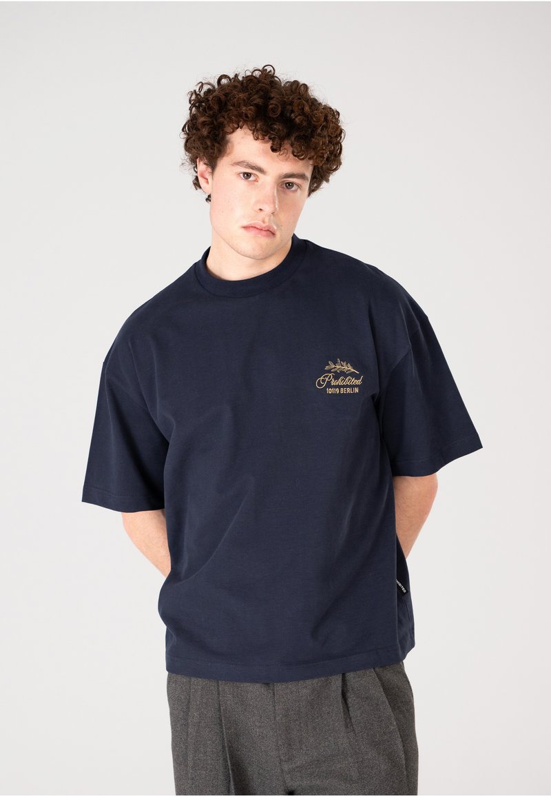 Camiseta de algodón azul marino con un corte relajado, mangas cortas y un logo impreso en dorado en el pecho. Tejido texturizado, diseño minimalista.