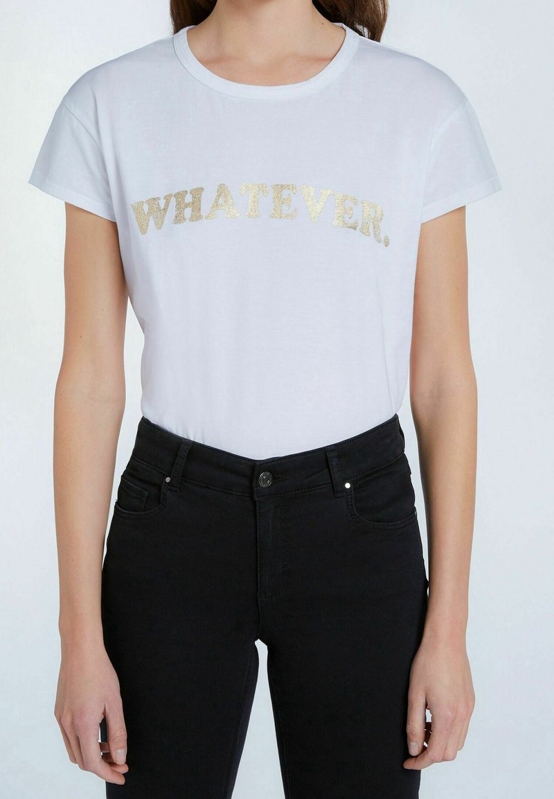 T-shirt en coton blanc à manches courtes avec un texte métallique doré indiquant "WHATEVER." Associé à un jean noir.