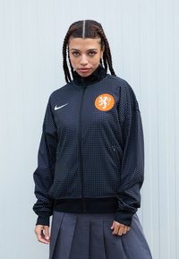 Nike bunda v černé barvě s bílým tečkovaným vzorem, vysokým límcem, zipovým zapínáním a oranžovým emblémem lva. Obsahuje postranní kapsy a žebrované manžety.