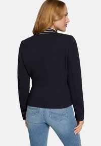 Veste ajustée bleu marine en tissu texturé, épaules structurées et une couture arrière détaillée, associée à un jean bleu clair.