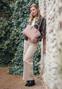Světle růžová taška přes rameno se strukturovaným designem, dvojitými uchy a zlatými akcenty. Modelka má na sobě tmavou koženou bundu a béžové kalhoty, stojí vedle kamenné zdi.
