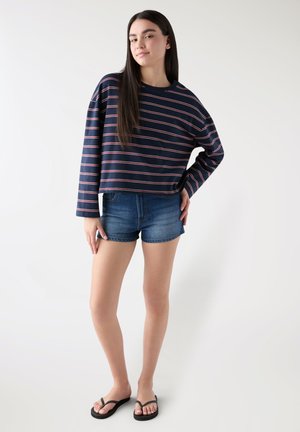 Giovane donna in piedi con la mano sul fianco, indossa una maglietta a maniche lunghe a righe blu navy, pantaloncini di jeans e infradito nere su sfondo bianco.