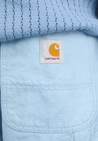 Tissu Carhartt bleu clair avec un écusson logo Carhartt jaune et blanc cousu sur une poche sous un tissu tricoté bleu texturé.