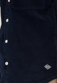 Marinblå kord skjorta med texturerat mönster, med en ficka med fickor och vita knappar. Inkluderar en liten logotyp vid fållen.