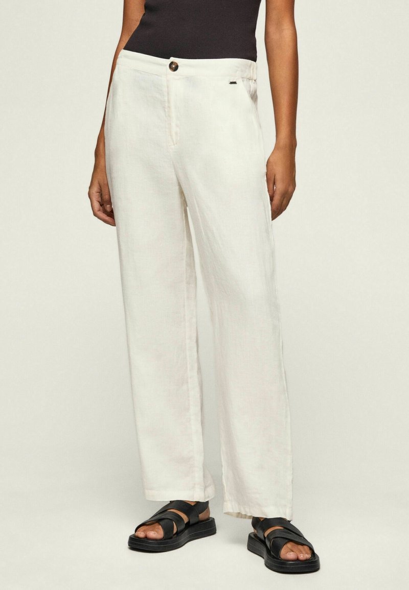 Personne portant un pantalon blanc ample, un haut noir sans manches et des sandales noires à brides, debout sur un fond clair uni.
