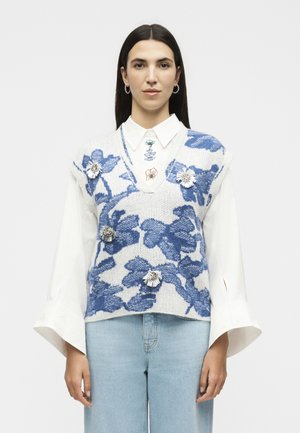 Kvinde med glat mørkt hår bærer en hvid skjorte under en blå og hvid blomsterstrikket ærmeløs sweater med blomsterbroscher og lyseblå jeans.