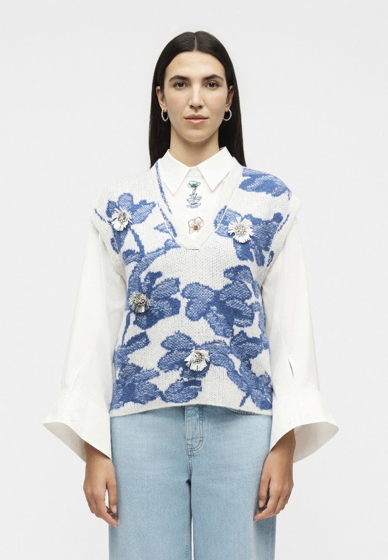 Femme aux cheveux raides et foncés porte une chemise blanche sous un pull sans manches tricoté bleu et blanc à fleurs avec des broches florales et un jean bleu clair.