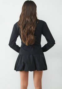 Schwarze Jacke mit langen Ärmeln, tailliertem Oberteil und ausgestellt rüschlichem Saum. Glattes Gewebe mit subtiler Glanzwirkung, von hinten gezeigt. Lange, wellige Haare sichtbar.