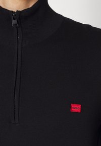 Schwarze Zip-Up-Sweatjacke mit hohem Kragen, gerippter Textur und einem kleinen roten rechteckigen Logo-Patch mit der Aufschrift "HUGO" auf der linken Seite.