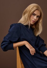 Plavolaska ženska z modrimi očmi, oblečena v mornarsko modro obleko, sedi na lesenem stolu pred enobarvno rjavim ozadjem.