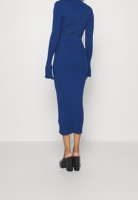 Abito blu a maglia con maniche lunghe, silhouette aderente, texture a costine, lunghezza al ginocchio e orlo scallop. Abbinato a tacchi alti neri.