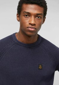 Giovane uomo con capelli corti e ricci che indossa un maglione lavorato a maglia blu navy con una piccola toppa gialla ricamata sul petto.