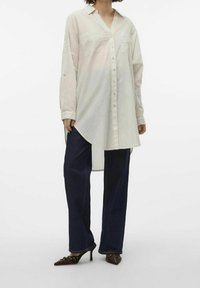 Donna che indossa una camicia beige lunga e morbida con strisce chiare, jeans larghi scuri e tacchi a punta marroni su uno sfondo bianco.