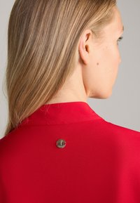 Rode blouse met een gladde textuur en hals, voorzien van een ronde gouden accent aan de achterkant met een bloemmotief en rode draad.