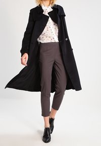 Cappotto trench nero doppiopetto con vita con cintura, indossato sopra una blusa floreale bianca e pantaloni grigi su misura. Stivaletti neri alla caviglia.