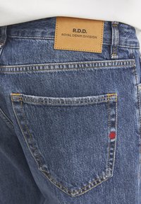 Tmavomodré denimové džínsy s oranžovým prešívaním, s hnedou koženou nášivkou s označením "R.D.D. ROYAL DENIM DIVISION" a malým červeným akcentom pri vrecku.