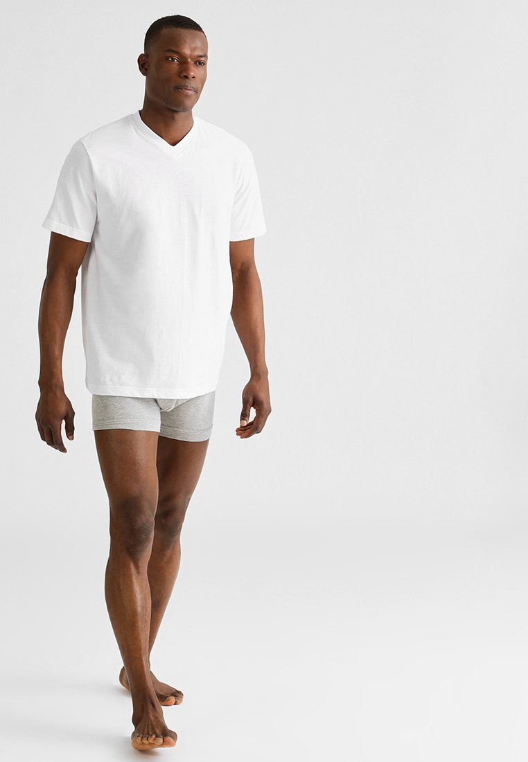 T-shirt blanc à col en V avec des manches courtes, associé à un short en boxeur gris, porté par une personne aux cheveux courts, debout pieds nus.