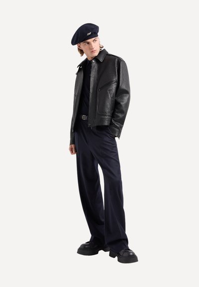 Giacca di pelle nera con texture, chiusura a zip, indossata sopra una camicia scura, abbinata a pantaloni larghi blu navy e scarpe nere robuste, completata da un berretto blu navy.