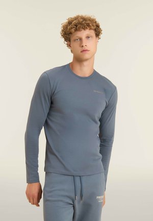 Giovane uomo con capelli ricci che indossa una maglia blu aderente a maniche lunghe e pantaloni della tuta abbinati, su uno sfondo chiaro e semplice.