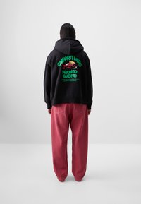 Black hoodie with green and red graphic text on the back, paired with loose-fitting, faded pink pants, featuring a clean, minimalistic design.