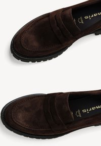 Bruna mockaläder loafers med en rektangulär detalj på framfoten, svart gummisula och synlig sömdetalj längs kanterna.