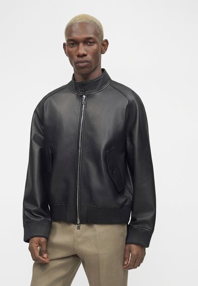 Μαύρο δερμάτινο μπουφάν με μπροστινό φερμουάρ και γιακά με κουμπιά, που φοριέται από άνδρα με κοντά ξανθά μαλλιά και μπεζ παντελόνια.