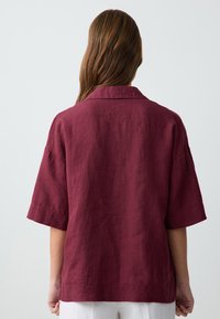 Camicia di lino bordeaux con una vestibilità rilassata, maniche corte e colletto classico. Texture liscia con fessure laterali all'orlo.