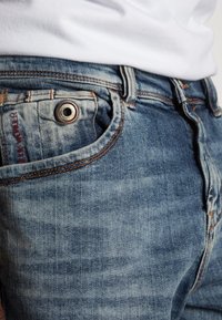 Närbild av en ficka på blåa jeans med en synlig knapp och en del av en vit t-shirt instoppad.