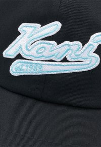 Must päikeklapp, millel on tikitud valge ja helesinise "Kani" logo ning aasta "1989" all. Tekstuuriga kangas ja kumer ääreosa.