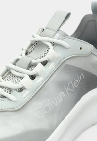 Ανοιχτό γκρι αθλητικό παπούτσι με λείο, γυαλιστερό επάνω μέρος και λεπτομέρειες από δίχτυ, λευκά κορδόνια και υφή στη σόλα. Το λογότυπο "Calvin Klein" είναι ανάγλυφο στο πλάι.