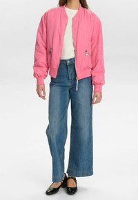 Růžová bomber bunda s elastickými manžetami, kombinovaná s modrými džíny s vysokým pasem a širokými nohavicemi a černými balerínkami. Texturovaná látka a viditelné zipy.
