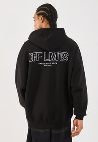 Person med fletter iført en svart hettegenser med teksten "OFF LIMITS DANGEROUS AREA BERLIN 2001" på ryggen mot en hvit bakgrunn.