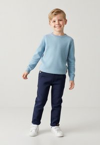 Jasnoniebieski sweter z dzianiny, granatowe spodnie z kieszeniami oraz białe tenisówki. Dziecko stoi na neutralnym tle, uśmiechając się, prezentując casualowy strój.