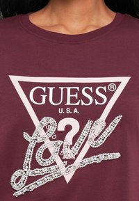 Close-up van een kastanjebruine shirt met een lichtroze driehoekig GUESS U.S.A. logo en de cursieve tekst "Love" versierd met parels en strass.