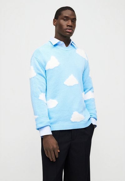 Νεαρός άνδρας που φοράει ένα ανοιχτό μπλε πουλόβερ με λευκά σύννεφα πάνω από πουκάμισο με γιακά, στέκεται με το ένα χέρι στην τσέπη μπροστά σε ένα απλό φόντο.