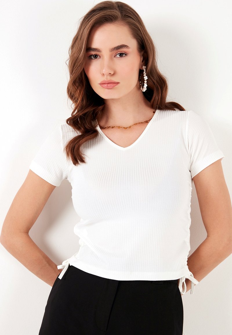 Top bianco a costine a maniche corte con scollatura a V, presenta lati con cordoncino per una vestibilità regolabile, abbinato a pantaloni neri.