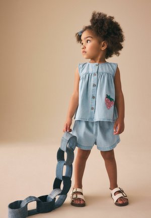 Ung pige med krøllet hår iført lys blå ærmeløs top og shorts, holder store kædelinks af denim, stående mod en ensfarvet beige baggrund.