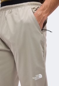 Κοντινή λήψη μπεζ παντελονιού North Face με ελαστική μέση και χέρι που βρίσκεται μερικώς στη πλαϊνή τσέπη σε απλό ανοιχτόχρωμο φόντο.