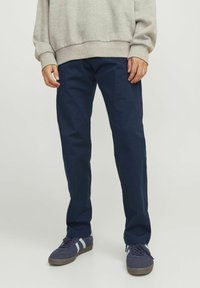 Person iført marineblå bukser, blå sneakers med hvide striber og en lysegrå sweatshirt, stående mod en ensfarvet baggrund.