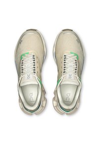 Beige athletic shoes with a textured upper, green accents, and white laces. Features a cushioned sole and logo on the tongue and side.