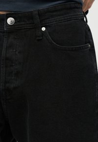 Κοντινή λήψη της περιοχής της μέσης μαύρων τζιν ντένιμ με εμπρόσθια τσέπη, θηλιά ζώνης και λεπτομέρεια ασημί κουμπιού, με φόντο ένα σκούρο πουκάμισο.