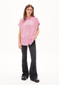 Pink short-sleeve button-up shirt with a relaxed fit, paired with black flared jeans and chunky black shoes, standing against a white background.